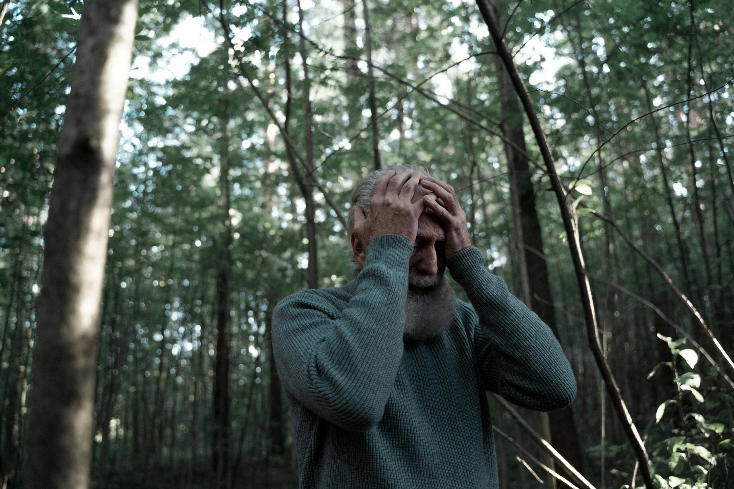 Homem idoso com as mãos na cabeça em meio à floresta, simbolizando esgotamento emocional e cansaço mental.